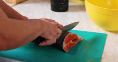 Ev mutfağında mavi bir kesme tahtasıyla somon balığını kesen kadın eli pek de yakın değil.