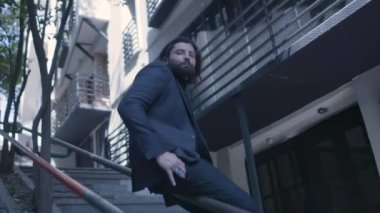 Takım elbiseli yakışıklı sakallı adam merdivenlerde ağır çekimde oturuyor.