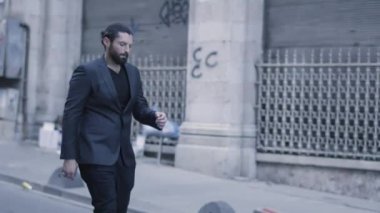 Takım elbiseli yakışıklı sakallı adam yavaş çekimde sokakta yürüyor.