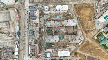 Geliştirmenin çeşitli aşamalarında çeşitli binaların yer aldığı büyük bir inşaat alanının havadan görünüşü, vinçler ve altyapı görülebilir.