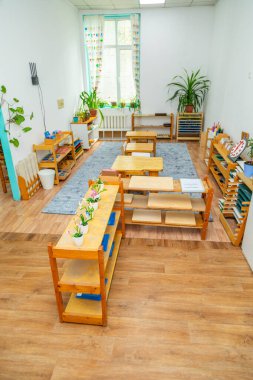 Montessori sınıfı ahşap mobilya, bitki ve eğitim malzemeleriyle dolu. Oda doğal ışıkla aydınlanmış..