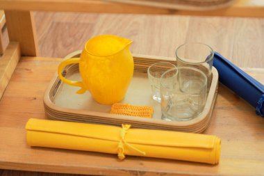 Montessori tray containing a yellow pitcher, glasses, and a cloth for teaching practical life skills and fine motor coordination in children clipart