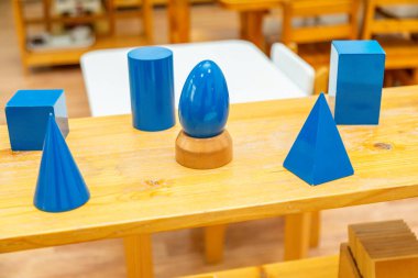 Montessori sınıfının rafında çocukların duyusal öğrenmesi için koni, küp ve silindirler gibi mavi geometrik katılarla ahşap tepsiler sergileniyor.