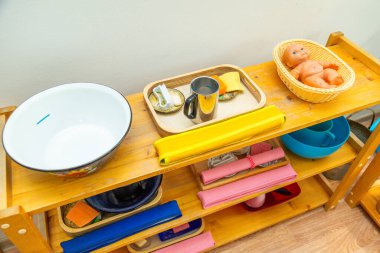 Tahta Montessori rafında sepetteki bir bebek, bir kap, sabun ve sünger gibi ev aletleriyle dolu bir tepsi ve pratik yaşam malzemesi var.