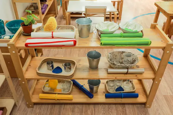 Erken eğitimde kullanılan tepsiler, kaplar, kovalar ve katlanmış kumaşlar gibi pratik yaşam malzemelerini gösteren ahşap bir Montessori rafı.