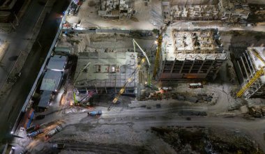 Geceleri vinçleri, beton pompaları ve ağır makineleri olan büyük bir bina inşa ediliyor. Arka planda sokak lambalarıyla aydınlatılmış kentsel konut binaları.