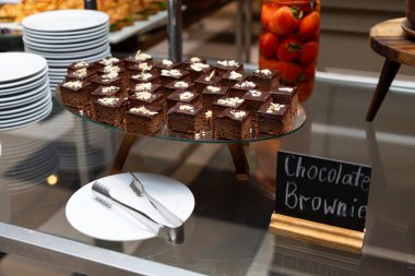 Sıralar dolusu çikolatalı browni kareleri cam bir standın üzerindeki badem dilimleriyle süslenmiş ve yakınlarda maşa ve tabaklar var.