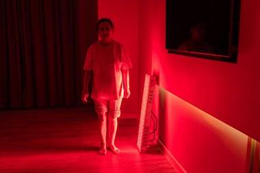 Evde kırmızı ışık terapi panelinin yanında duran yaşlı bir kadın.