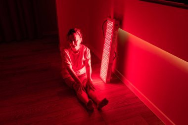 Yerde bacakları kırmızı ışık terapi cihazının yanında uzanmış yaşlı bir kadın oturuyor.