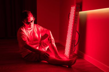 Yaşlı kadın içeride bacağında kırmızı ışık terapi paneli kullanarak oturuyor.