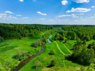 Geniş bir hava manzarası, açık mavi gökyüzünün altında sık ormanlarla çevrili yeşil bir vadiden geçen kıvrımlı bir nehri ortaya çıkarıyor.