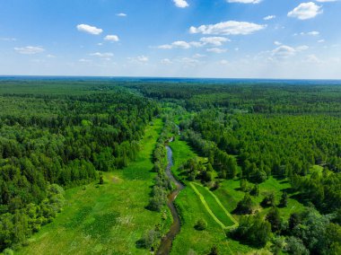 Geniş yeşil ormanlar ve tarlalar arasında dolanan geniş ve dolambaçlı bir nehri, açık mavi gökyüzünün altında dağınık bulutlarla gösteren geniş bir hava görüntüsü vardır.