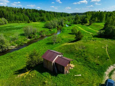 Havadan bakıldığında, kıvrımlı bir nehrin kenarında, yemyeşil tarlalar ve parlak bir gökyüzünün altındaki sık ormanlarla çevrili bir kır evi görülüyor.