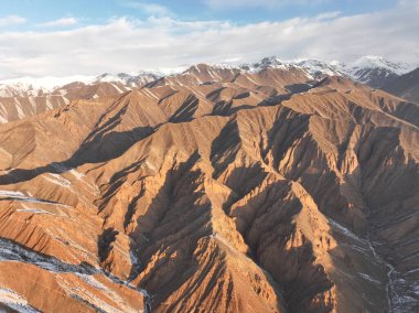 Geniş, engebeli, kahverengi dağ sıraları, açık mavi gökyüzünün altında güneş tarafından güzelce aydınlatılan kar parçaları.