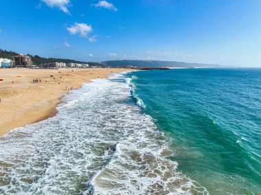 Geniş bir kumsalda ilerleyen açık mavi dalga ve yakınlarda yürüyen birkaç kişi.