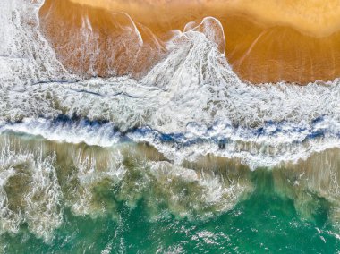 Kıyı dalgaları kıvrılıyor ve kumlu bir plaja dökülüyor görünür bir sörf köpüğüyle