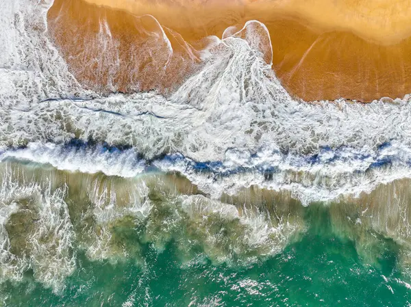 Kıyı dalgaları kıvrılıyor ve kumlu bir plaja dökülüyor görünür bir sörf köpüğüyle