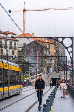 Porto, Portekiz - 12 Mayıs 2024: Çeşitli etnik kökenlere ve yaşlara sahip insanlar Dom Luis Köprüsü 'nün üst güvertesinde arka planda Porto' nun binalarının bulunduğu metro treninin yanında yürüyüp motosiklet kullanıyorlar.