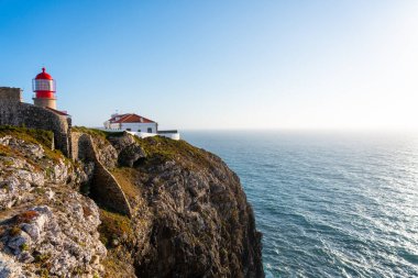 Cabo da Roca 'nın ikonik kırmızı ve beyaz deniz feneri engebeli bir uçurumun üzerinde berrak bir gökyüzünün altındaki mavi Atlantik Okyanusuna bakıyor.