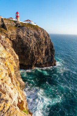 Kırmızı ve beyaz Cabo da Roca deniz feneri, dalgaların tabanına çarptığı canlı turkuaz ve mavi Atlantik Okyanusuna bakan dramatik bir uçurum kenarında duruyor.