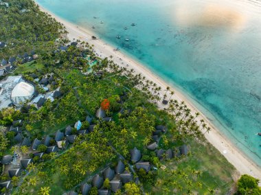 Gün batımında sahil manzaralı, tekneler, bungalovlar ve sahil boyunca uzanan palmiye ağaçları.