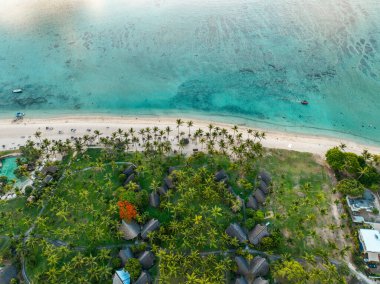 Gün batımında sahil manzaralı, tekneler, bungalovlar ve sahil boyunca uzanan palmiye ağaçları.