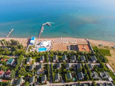 Rıhtım, yüzme havuzu, plaj şemsiyeleri ve ön planda kulübeleri olan bir tatil köyü sahili görüntüsü.