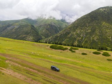 Geniş yeşil bir dağ vadisinde arka planda bulutlu tepeler olan toprak bir yolda giden siyah bir SUV 'nin insansız hava aracı görüntüsü.