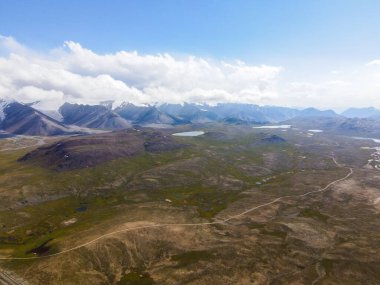 Dağılmış göller, sulak alanlar, toprak bir yol ve uzaktaki karla kaplı zirvelerle uzak bir dağ manzarası.