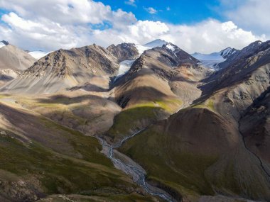 Engebeli zirveleri, buzulları ve bulutlu gökyüzünün altındaki vadiden akan bir nehrin dramatik bir dağ manzarasının insansız hava aracı görüntüsü.