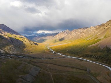 Geniş bir dağ vadisinde dolambaçlı bir nehrin drone görüntüsü dramatik ışık, fırtınalı bulutlar ve engebeli yamaçlar.