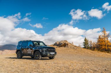Aktash, Altai Cumhuriyeti - 12 Ekim 2023: Siyah bir Jetour T2 SUV, sonbahar ağaçları ve bulutlu açık mavi gökyüzünün altındaki büyük kayalıklarla tozlu kahverengi bir tepenin üzerine park edilmiştir.