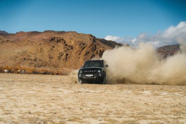Aktash, Altai Cumhuriyeti - 12 Ekim 2023: Siyah bir Jetour T2 SUV hızla off-road sürüşü yaparak, arka planda çorak tepeler ve bazı develerle büyük bir toz bulutu oluşturdu.
