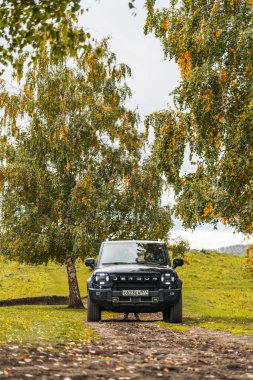 Aktash, Altai Cumhuriyeti - 12 Ekim 2023: Sarı ve yeşil sonbahar yapraklı iki huş ağacı arasında dar bir toprak yolda giden siyah bir Jetour T2 SUV 'nin ön görüntüsü