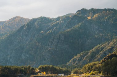 Yeşil ve sarı sonbahar ağaçlarının yoğun karışımıyla kaplı sarp bir dağın tabanda küçük bir kulübesi olan güzel bir manzara görüntüsü.