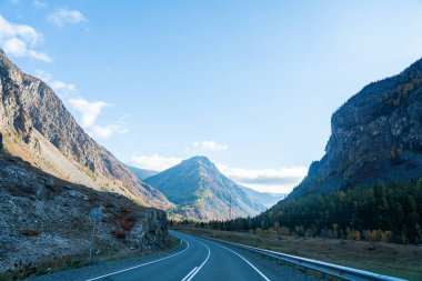 Uçsuz bucaksız, dolambaçlı bir yola açılan geniş, panoramik bir manzara. Parlak mavi gökyüzünün altındaki sonbahar ağaçlarıyla kaplı bir vadiye giden bir yol.