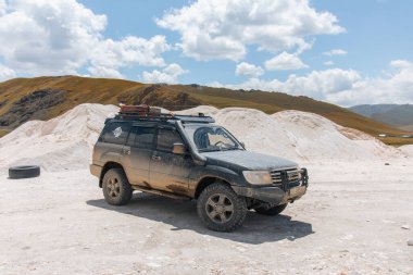 Naryn, Kırgızistan - 12 Ekim 2024: Karanlık bir off-road aracı bulutlu mavi gökyüzünün altında, beyaz mineral zengini bir tümseğin yanında yüksek irtifa yolunda duruyor.