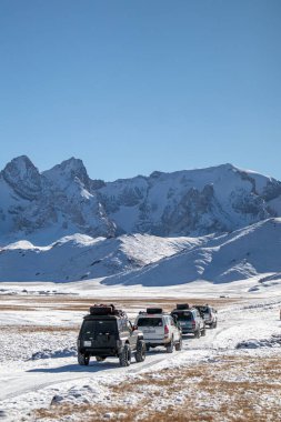 Naryn, Kırgızistan - 12 Ekim 2024: Bir off-road aracı konvoyu, arka planda görkemli dağların bulunduğu geniş bir vadide karla kaplı toprak bir yol boyunca ilerliyor