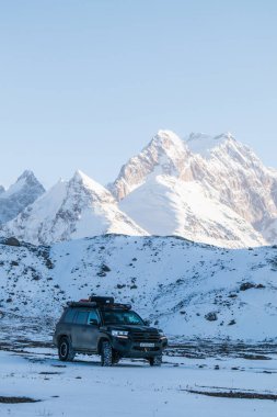 Naryn, Kırgızistan - 12 Ekim 2024: Karanlık bir off-road aracı soğuk bir kış gününde arka planında görkemli karlı bir dağ bulunan karla kaplı bir arazide duruyor