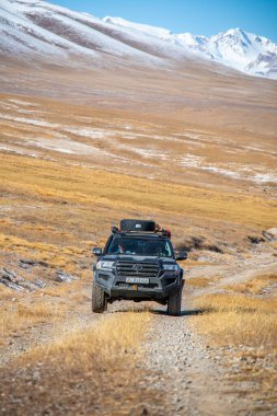 Naryn, Kırgızistan - 12 Ekim 2024: Toyota Land Cruiser arka planda karla kaplı bir dağ silsilesiyle uçsuz bucaksız, altın ve kahverengi bir vadide toprak bir yolda ilerliyor