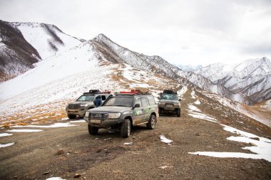 Naryn, Kırgızistan - 12 Ekim 2024: Bir off-road aracı konvoyu, arka planda karlı dağ sıraları olan geniş bir vadide, karlı bir dağ yolunda ilerliyor.