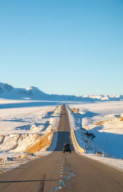 Naryn, Kırgızistan - 12 Ekim 2024: Karanlık bir off-road aracı, arka planda görkemli dağlarla kaplı engin karla kaplı bir vadide uzun ve dolambaçlı bir yolda ilerliyor.