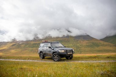 Naryn, Kırgızistan - 12 Ekim 2024: Karanlık bir off-road aracı engin, yeşil ve altın vadide uzanan görkemli dağları ve alçak bulutları olan