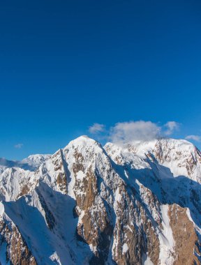 Berrak mavi gökyüzünün altında engebeli, kayalık, karlı dağların dikey görüntüsü.