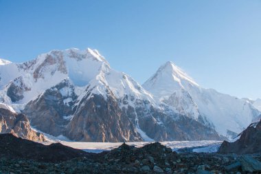 Karla kaplı görkemli bir dağ zirvesi engebeli, kayalık bir araziden yükseliyor ve berrak mavi bir gökyüzünün altında vadiden aşağı bir buzul akıyor.