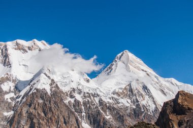 İki engebeli, karlı dağ zirvesinin yakın plan çekimi. Etrafında bulutlar uçuşuyor. Açık mavi bir gökyüzünün altında.