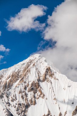 Beyaz bulutlu, mavi gökyüzünün altında engebeli, kayalık yamaçları olan görkemli, karlı bir dağın dikey yakın çekimi.