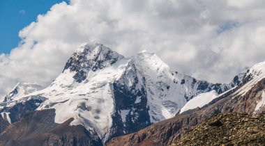 Arkaplanda kayalık bir ön planda ve dramatik bulutlu gökyüzü olan devasa, engebeli, karlı bir dağ sırasının panoramik görüntüsü.
