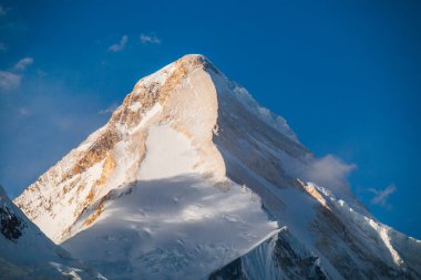 Görkemli bir karlı dağ zirvesi bir tarafı güneş ışığıyla aydınlanır, diğer tarafı ise açık mavi gökyüzünün altında gölgelerle kaplıdır.