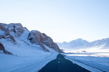 Uzun düz bir asfalt yol, her iki taraftaki karla kaplı dağlarla çevrili bir vadiden geçer. Açık mavi gökyüzünün altında.